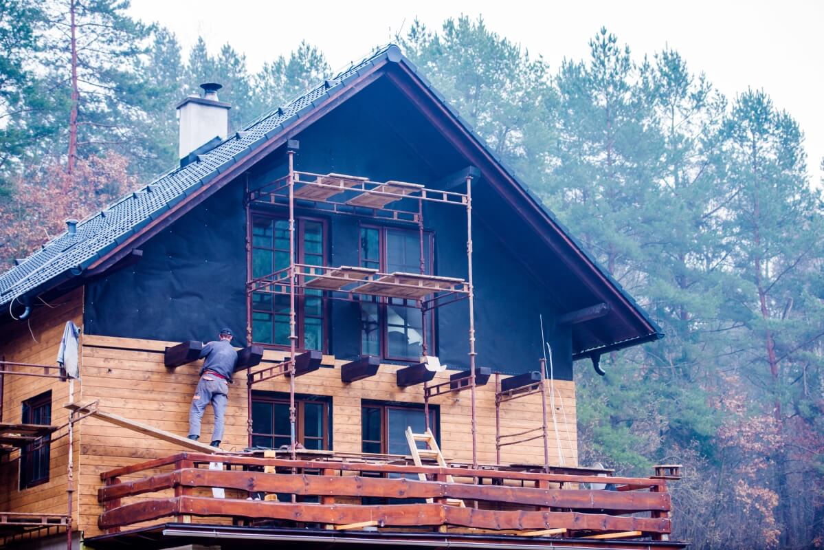 Robotnik pracuje na rusztowaniu przy elewacjach drewnianych domu w otoczeniu lasu.