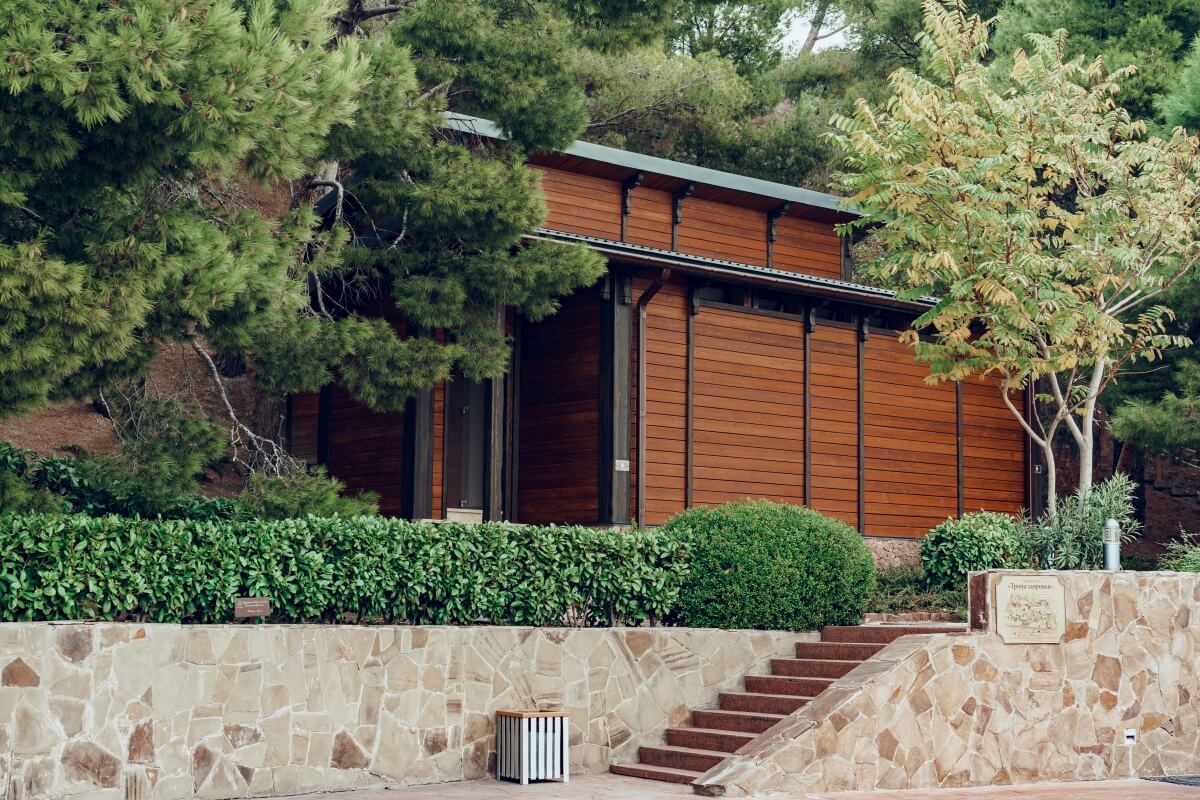 Drewniany budynek otoczony roślinnością, z szalówką elewacyjną malowaną na brązowo.